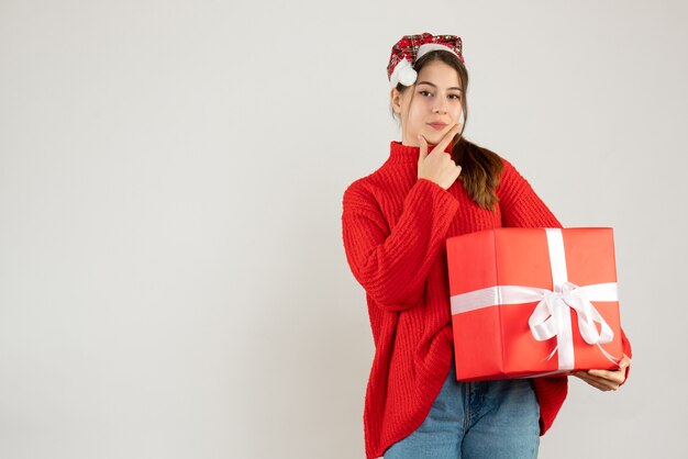 Feliz linda chica con gorro de Papá Noel con regalo poniendo el dedo en la barbilla de pie sobre blanco