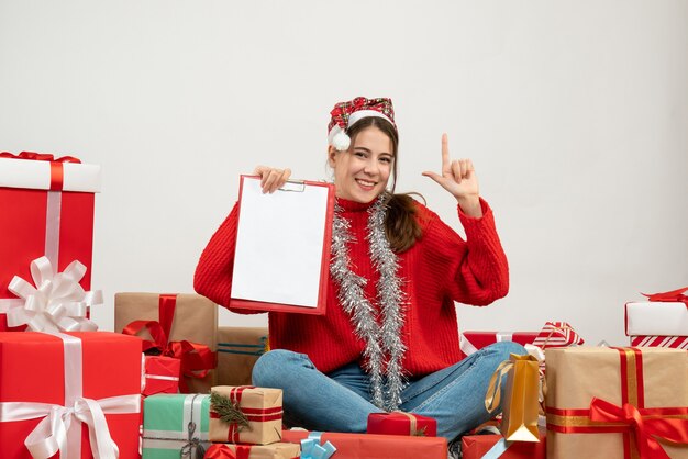 Feliz linda chica con gorro de Papá Noel sosteniendo el documento poniendo su pistola de dedo sentado alrededor presenta en blanco