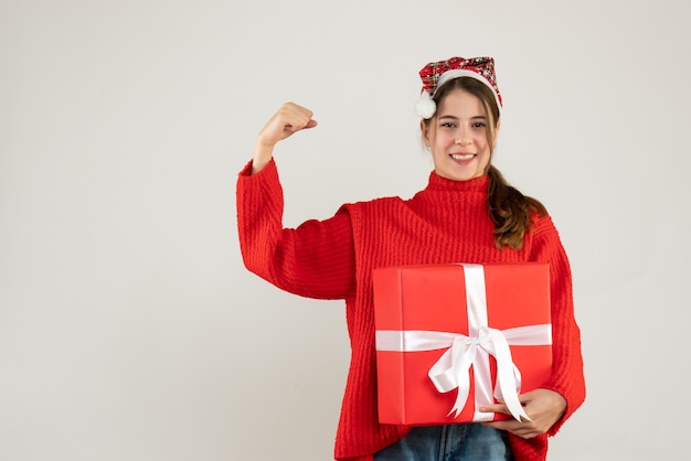 Feliz linda chica con gorro de Papá Noel sosteniendo presente mostrando gesto ganador de pie sobre blanco