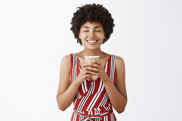 Feliz y linda joven afroamericana con peinado rizado, sosteniendo un vaso de papel con ambas manos, cerrando los ojos y sonriendo de satisfacción