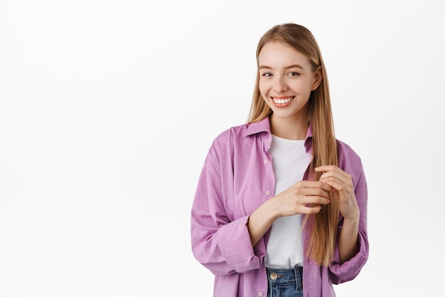 Feliz linda mujer rubia tocando su cabello liso y saludable sonriendo dientes blancos y mirando a la cámara de pie en ropa de verano de moda contra el fondo blanco