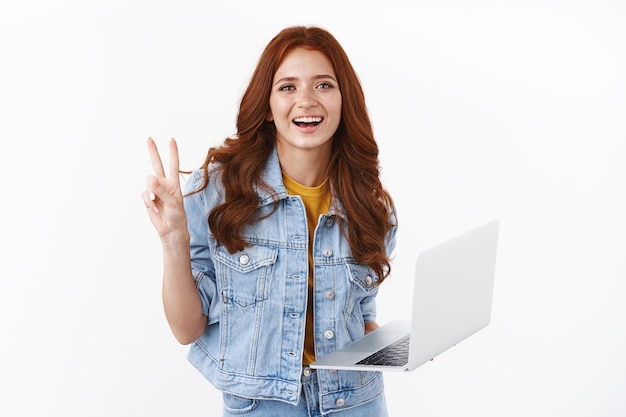 Feliz linda pelirroja independiente en chaqueta de mezclilla logra fácilmente la meta usando la computadora portátil, muestra la victoria de la paz, muestra de buena voluntad, sonriendo con alegría, trabajando en el control remoto, preparar el proyecto, pared blanca