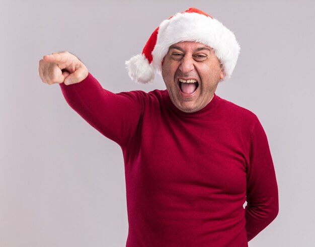 Feliz loco hombre de mediana edad vestido con gorro de Papá Noel de Navidad apuntando con el dedo índice a algo parado sobre la pared blanca