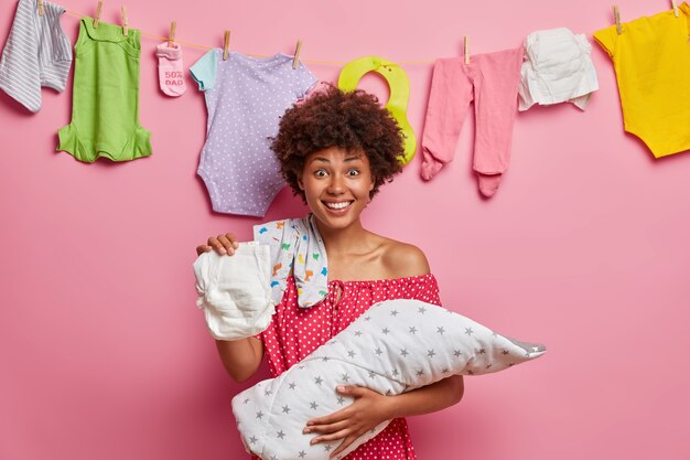 Feliz madre afroamericana se preocupa por el bebé, cambia el pañal a la hija, juega con el niño, se para en el interior de la casa, posa