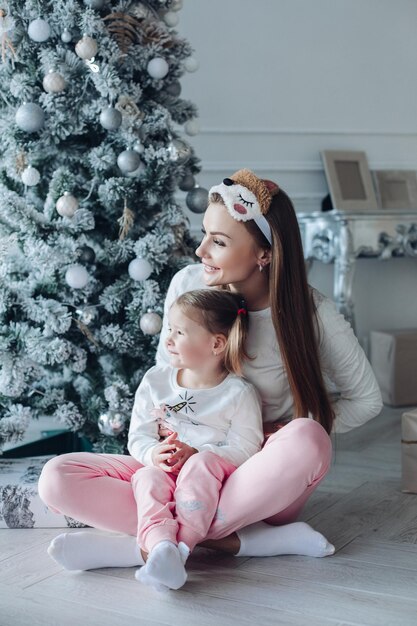 Feliz madre e hija festiva abrazándose sentados juntos en la decoración del hermoso árbol de navidad