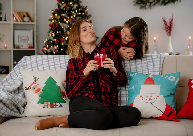 Feliz madre e hija mirándose la madre sostiene una caja de regalo sentada en el sofá y disfrutando de la Navidad en casa