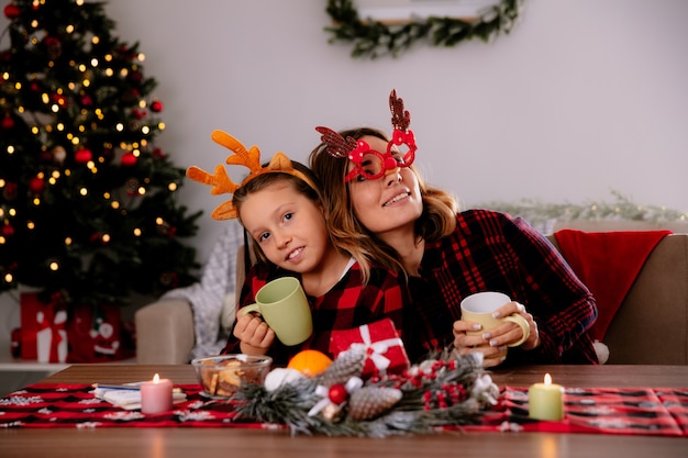 Feliz madre e hija sosteniendo la taza sentados a la mesa disfrutando de la Navidad en casa