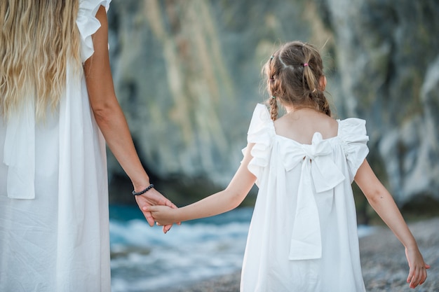 Feliz madre e hija en vestido blanco de pie y tomados de la mano en la orilla del mar durante la puesta de sol.