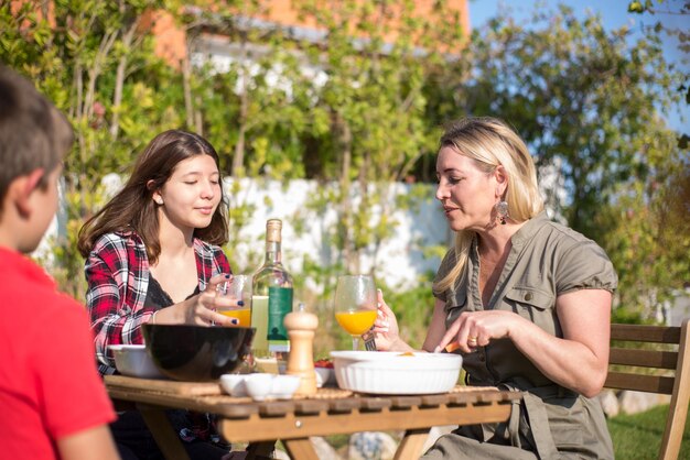 Feliz madre e hijos comiendo barbacoa en el patio trasero. Mujer adulta media y niños sentados alrededor de la mesa, comiendo comida recién cocinada, bebiendo bebidas. Barbacoa, cocina, comida, concepto familiar.