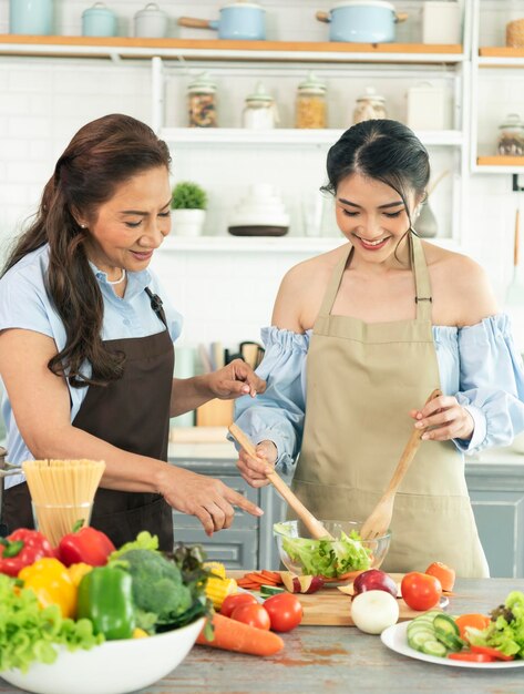 Feliz madre de familia asiática enseñando a su hija a hacer ensalada en la cocina en casa