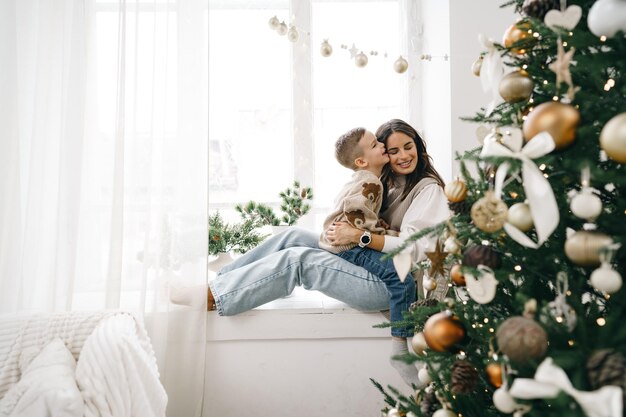 Feliz madre con hijo sentarse en el alféizar de la ventana cerca del árbol de Navidad