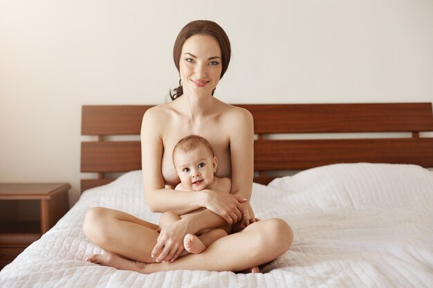 Feliz madre joven desnuda sonriendo abrazando a su bebé recién nacido agradable sentado en la cama juntos.