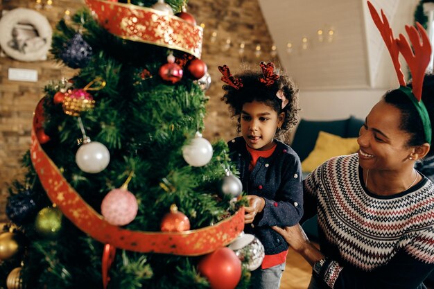 Feliz madre negra e hija decorando el árbol de Navidad en la sala de estar