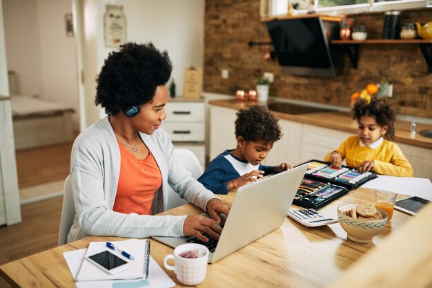 Feliz madre negra que trabaja usando una laptop mientras sus hijos colorean en casa