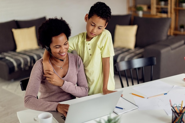 Feliz madre negra trabajando en una laptop mientras su hijo mira lo que está haciendo