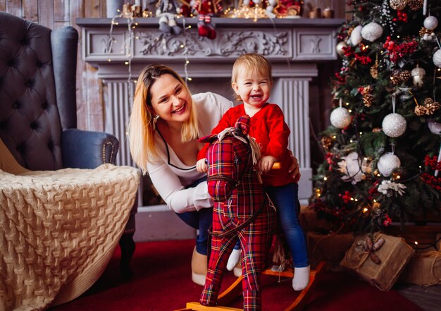 Feliz madre y niño encantador en el caballo de juguete posan ante un árbol de Navidad