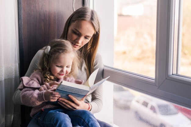 Feliz madre y pequeña hija leyendo libro, sentado en un cómodo sofá en la sala de estar