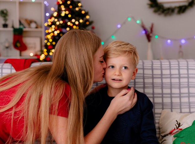 Feliz madre en vestido rojo besando a su pequeño hijo sentado en un sofá en una habitación decorada con árbol de Navidad en el fondo