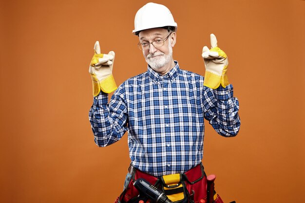 Feliz maduro trabajador de la construcción masculino jubilado con guantes de goma amarillos, riñonera y casco blanco mirando con una amplia sonrisa alegre, listo para trabajar, apuntando con ambos dedos índices hacia arriba