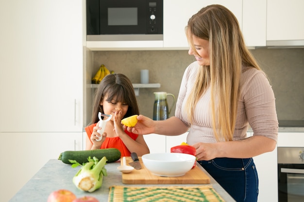 Feliz mamá e hija divirtiéndose mientras preparan la cena juntos. Niña y su madre pelando y cortando verduras para ensalada y limón para vestirse en la cocina. Concepto de cocina familiar