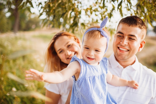 Feliz mamá y papá sostienen a pequeña hija en sus brazos de pie en los rayos del sol de la tarde