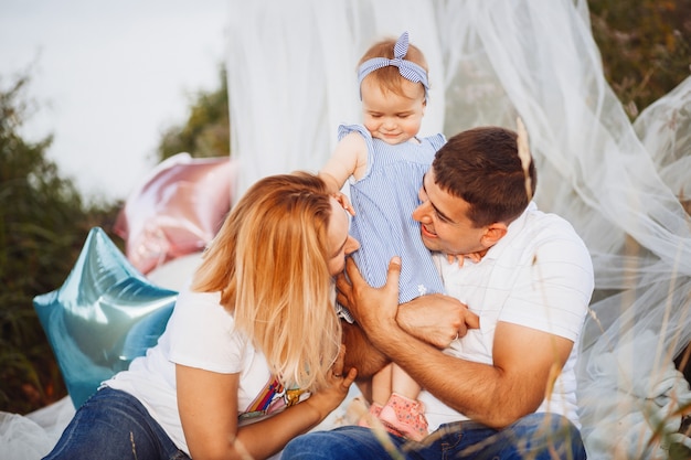 Feliz mamá y papá sostienen a la pequeña hija en sus brazos sentados debajo de la tienda blanca