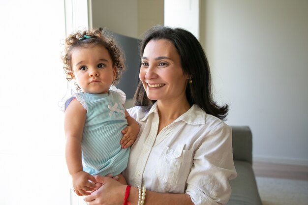 Feliz mamá de pelo negro con dulce hija en brazos. Linda niña de pelo rizado mirando a otro lado. Tiro medio. Concepto de paternidad e infancia