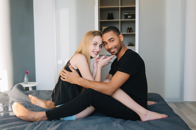 Feliz mañana juntos en casa de chico guapo y mujer joven con largo cabello rubio sonriendo. Relajarse en la cama en un apartamento moderno, relación, emociones verdaderas, enamorado