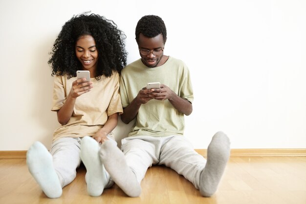 Feliz matrimonio joven descansando en casa sobre un piso de madera con gadgets. Chica guapa enviando mensajes de texto a amigos en línea a través de las redes sociales mientras su esposo está sentado a su lado