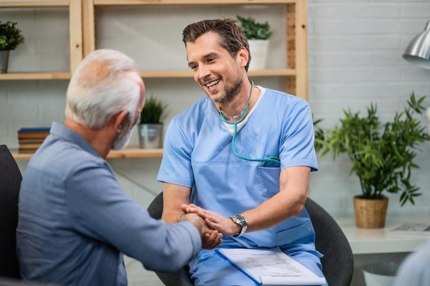 Feliz médico general hablando con un anciano mientras le da la mano durante una visita a casa