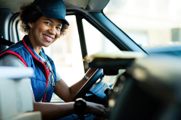 Feliz mensajera negra conduciendo una furgoneta de reparto y mirando a la cámara