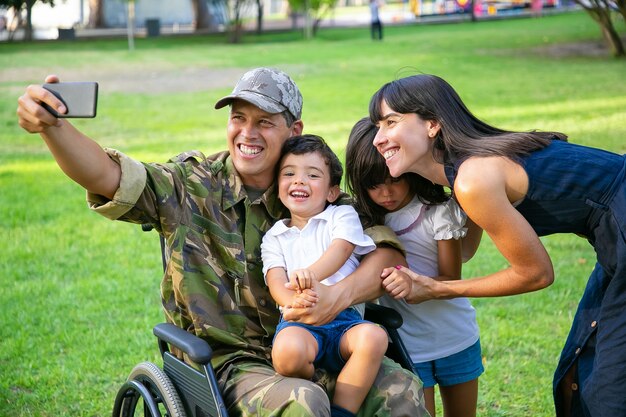 Feliz militar discapacitado tomando selfie con su esposa y sus dos hijos en el parque. Veterano de guerra o tiempo libre con concepto de familia.