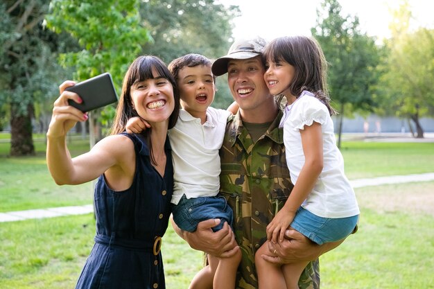 Feliz militar emocionado, su esposa y dos hijos celebrando el regreso de los papás, disfrutando del tiempo libre en el parque, tomando selfie en el teléfono celular. Tiro medio. Reunión familiar o concepto de regreso a casa