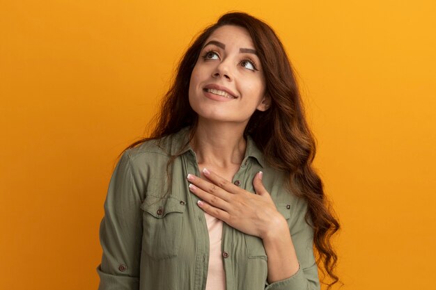 Feliz mirando ap joven hermosa chica con camiseta verde oliva poniendo la mano en el corazón aislado en la pared amarilla