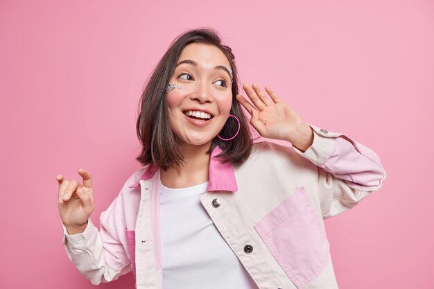 Feliz modelo de mujer asiática bastante joven levanta la mano en alto disfruta de libertad baila sonrisas despreocupadas concentradas positivamente vestida con ropa elegante se siente feliz aislado sobre pared rosa.