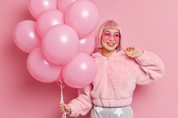 Feliz modelo de mujer asiática de buen aspecto con cabello rosado vestida con abrigo de piel de invierno lleva gafas de sol en forma de corazón de moda sostiene globos se prepara para la celebración de una ocasión especial. Concepto de evento festivo