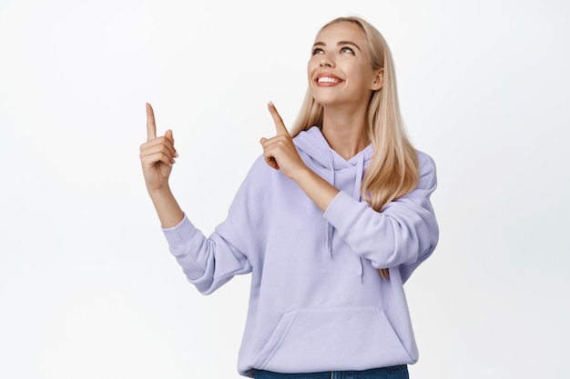 Feliz modelo de mujer bonita con cabello rubio señalando con el dedo y mirando la esquina superior izquierda leyendo el logotipo o pancarta con una sonrisa complacida de fondo blanco