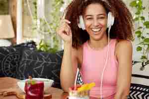 Foto gratuita feliz modelo de mujer de piel oscura con amplia sonrisa, se ríe alegremente mientras escucha anécdotas en línea en auriculares, conectado a un teléfono inteligente moderno, publica una publicación de internet en el sitio web, descansa en el café.