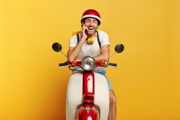 Feliz motociclista posa en su propio transporte rápido, llama al cliente a través del teléfono inteligente, viaja a larga distancia, usa casco, auriculares estéreo alrededor del cuello, sonríe a la cámara. Conductor masculino conduce scooter