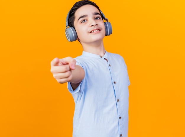 Feliz muchacho caucásico joven con auriculares mirando y apuntando a la cámara aislada sobre fondo naranja con espacio de copia