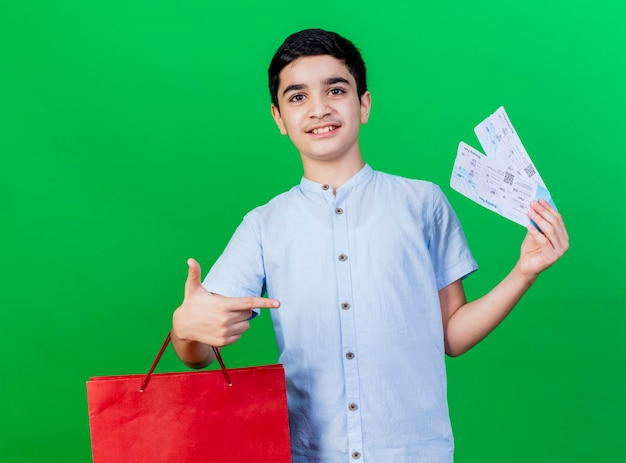 Feliz muchacho caucásico joven con bolsa de compras y billetes de avión apuntando a billetes mirando a cámara aislada sobre fondo verde