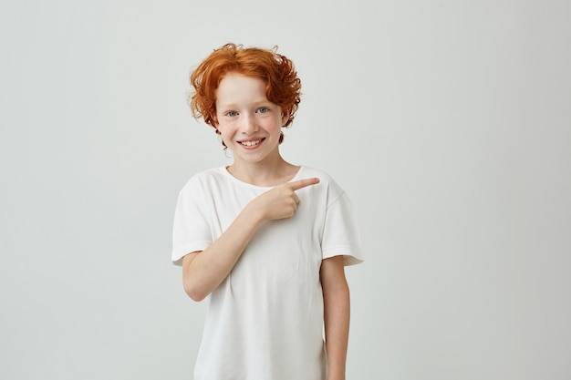 Feliz muchacho lindo sonriente con el pelo rizado rojo y pecas apuntando con el dedo a un lado con expresión alegre.