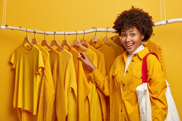 Feliz mujer adicta a las compras elige ropa en perchas en su propio armario, vestida con chaqueta brillante, lleva bolso, tiene una sonrisa atractiva