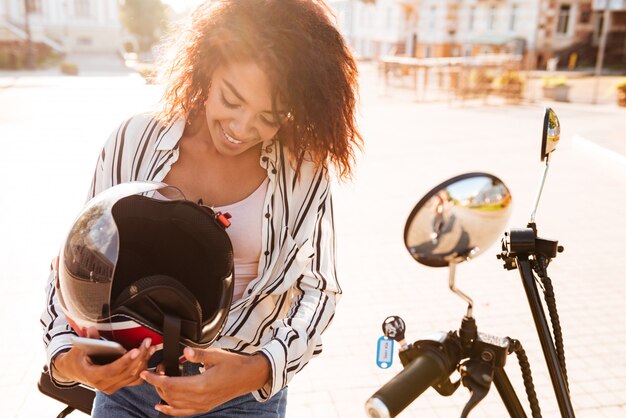 Feliz mujer africana sentada en moto moderna al aire libre y sosteniendo halmet