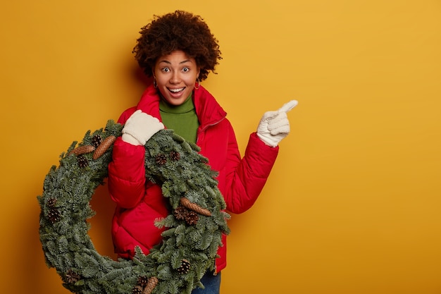 Feliz mujer afro muestra el camino a su casa, viste abrigo rojo, guantes blancos, lleva corona de navidad, apunta al espacio en blanco, se destaca sobre fondo amarillo