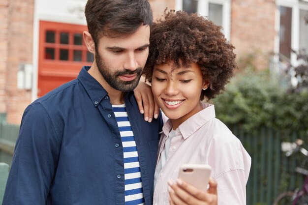 Feliz mujer afroamericana muestra video online de redes sociales en celular a su novio barbudo