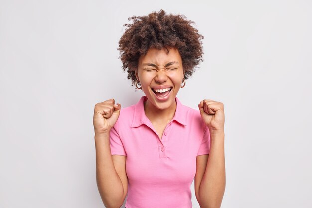 Feliz mujer afroamericana de pelo rizado hace bomba de puño se regocija con los resultados positivos viste una camiseta rosa informal