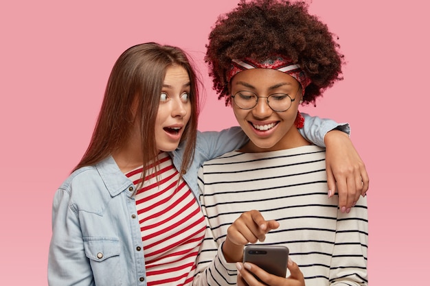 Foto gratuita feliz mujer afroamericana de piel oscura con corte de pelo afro mira felizmente al teléfono inteligente, sonríe con alegría, sostiene el teléfono inteligente