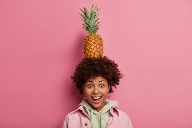 Feliz mujer afroamericana sostiene deliciosa piña madura fresca en la cabeza, mira con alegría a la cámara, se divierte solo con frutas tropicales, tiene buen humor