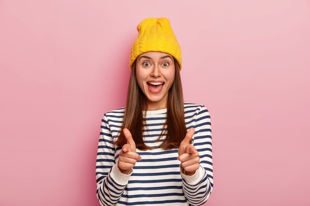 Feliz mujer apunta directamente a la cámara, expresa su elección con un gesto de pistola, viste un sombrero amarillo y un jersey informal a rayas